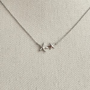 May include: A silver-toned necklace featuring a delicate chain and two attached starfish charms of different sizes. The larger starfish is positioned to the left of the smaller one, both suspended horizontally along the center of the thin metal chain.