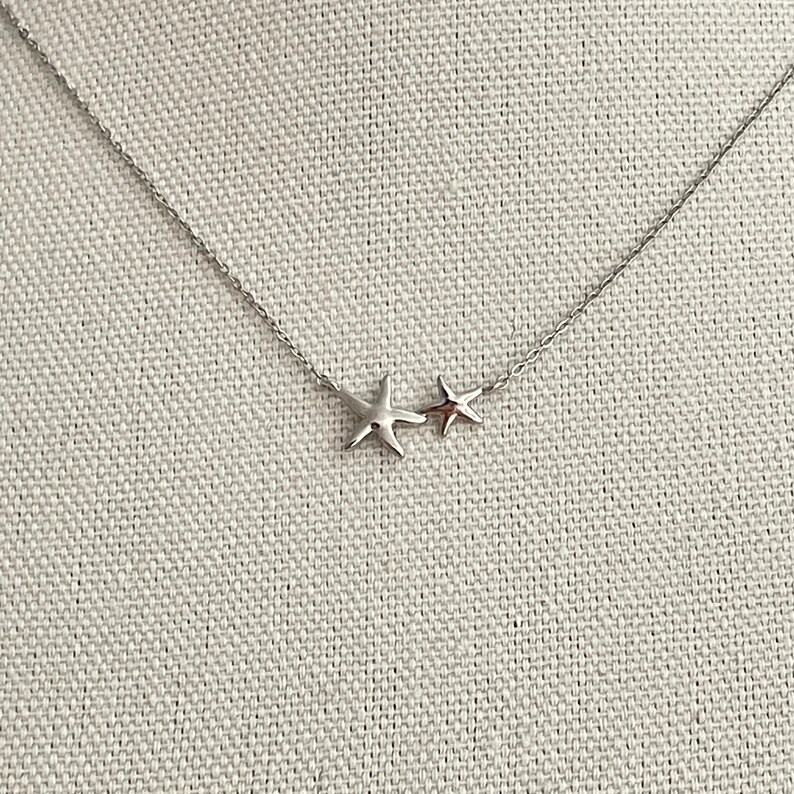 May include: A silver-toned necklace featuring a delicate chain and two attached starfish charms of different sizes. The larger starfish is positioned to the left of the smaller one, both suspended horizontally along the center of the thin metal chain.