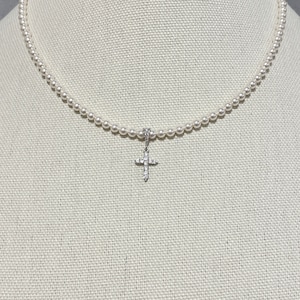 May include: A white pearl necklace with a small, silver cross pendant encrusted with clear rhinestones. The necklace is displayed on a white mannequin.