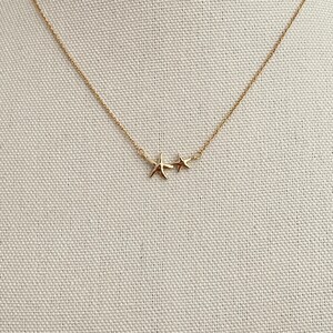 May include: A gold-toned necklace featuring a delicate chain with two attached charms, one shaped like a five-pointed starfish and the other a smaller five-pointed star, both positioned centrally along the thin metallic link chain.