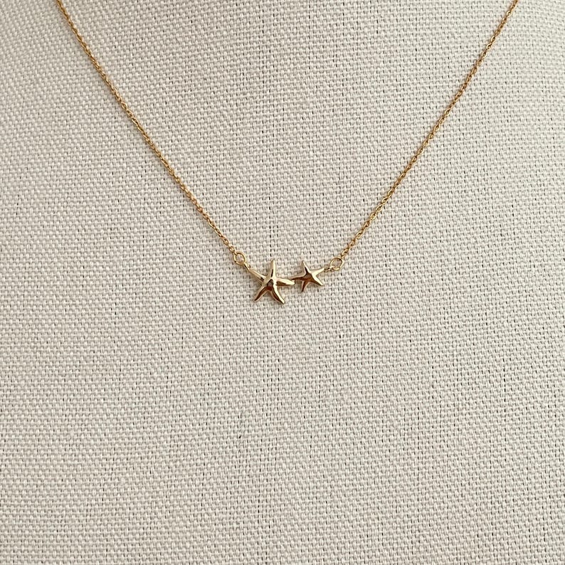 May include: A gold-toned necklace featuring a delicate chain with two attached charms, one shaped like a five-pointed starfish and the other a smaller five-pointed star, both positioned centrally along the thin metallic link chain.