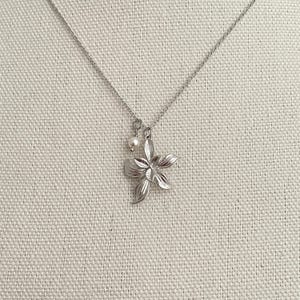May include: A silver-toned necklace featuring a flower pendant with detailed petals and a small pearl charm. The chain is delicate, and the pendant is centered against a neutral background, showcasing the jewelry's design.