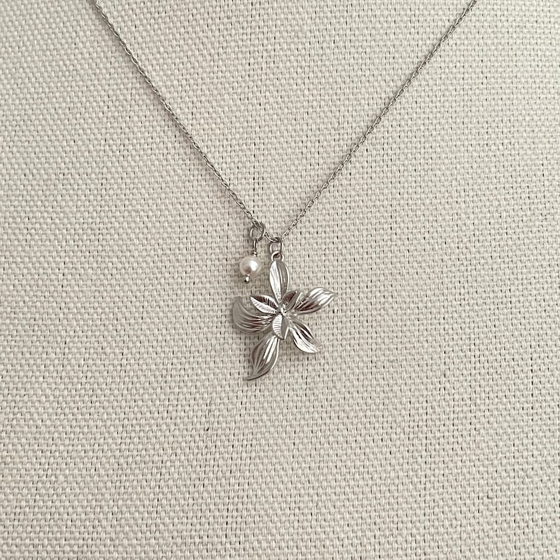 May include: A silver-toned necklace featuring a flower pendant with detailed petals and a small pearl charm. The chain is delicate, and the pendant is centered against a neutral background, showcasing the jewelry's design.