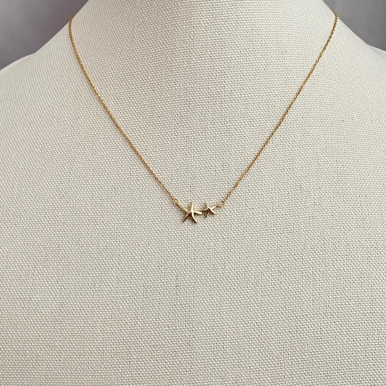 May include: A gold-toned necklace featuring a delicate chain and two small starfish charms positioned off-center. The metal has a polished finish, and the two starfish are of slightly different sizes, creating a simple and cohesive design.