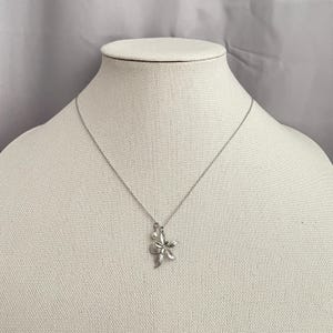 May include: A silver-toned necklace with a flower pendant and a small pearl. The flower pendant has detailed petals. The necklace is displayed on a white mannequin.