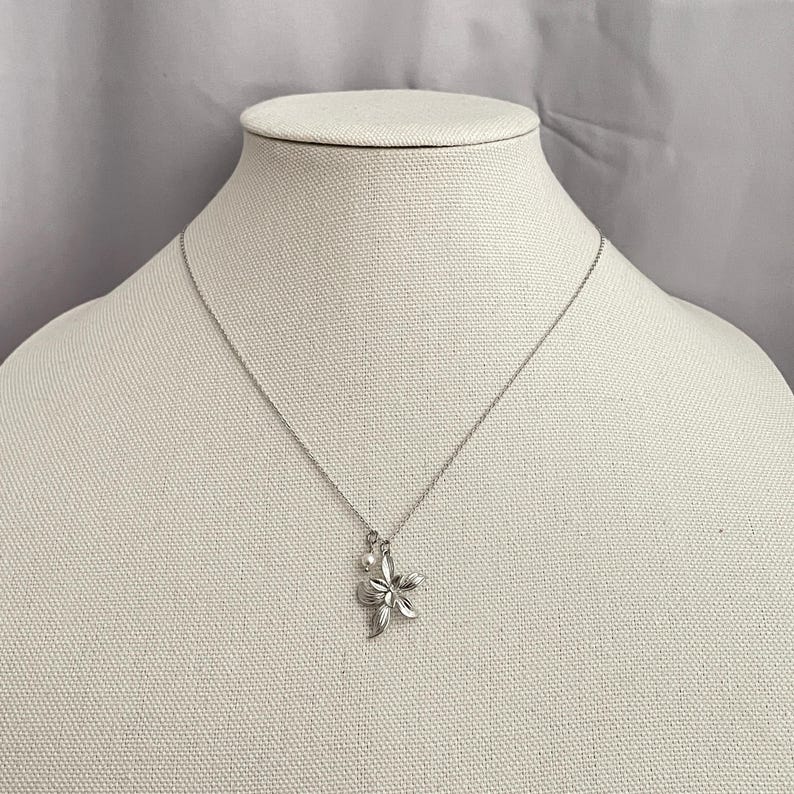 May include: A silver-toned necklace with a flower pendant and a small pearl. The flower pendant has detailed petals. The necklace is displayed on a white mannequin.