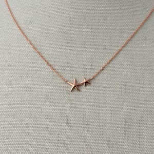 May include: A rose gold necklace featuring a delicate chain and two small starfish charms. The larger starfish is positioned slightly to the left of the smaller one, both suspended from the thin, metallic rose gold link chain.