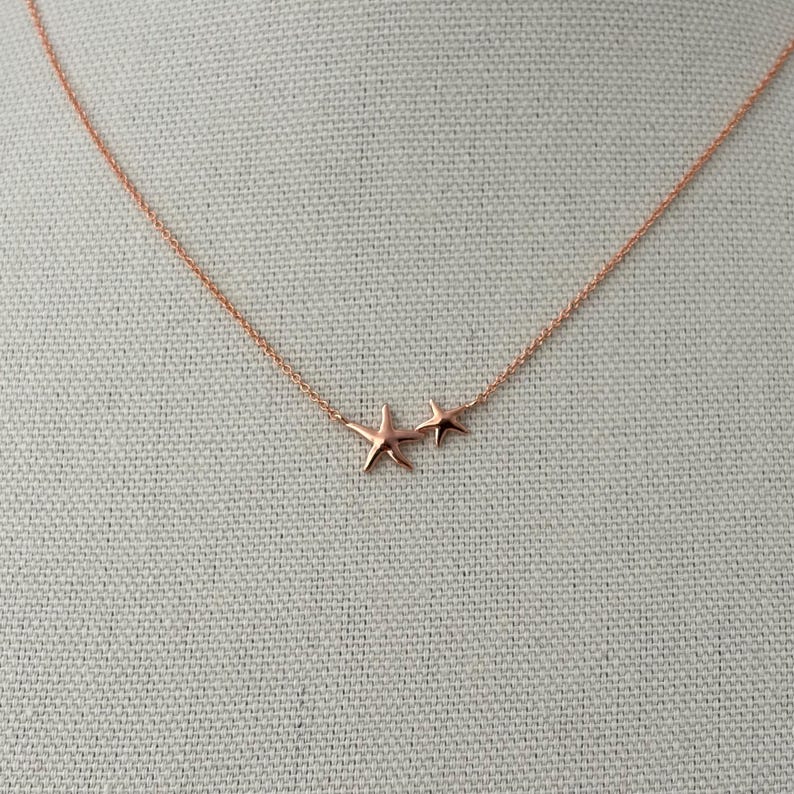 May include: A rose gold necklace featuring a delicate chain and two small starfish charms. The larger starfish is positioned slightly to the left of the smaller one, both suspended from the thin, metallic rose gold link chain.