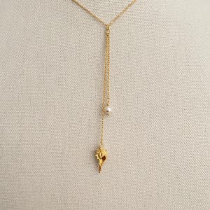 May include: A gold-colored necklace with a delicate chain. The necklace features a small, white pearl and a seashell pendant. The seashell pendant is intricately detailed. The necklace is displayed against a neutral background.