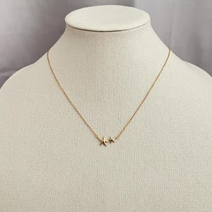 May include: A gold-toned necklace featuring a delicate chain and two small starfish-shaped pendants positioned together. The metal has a polished finish, and the design includes one larger starfish charm next to a slightly smaller starfish charm.