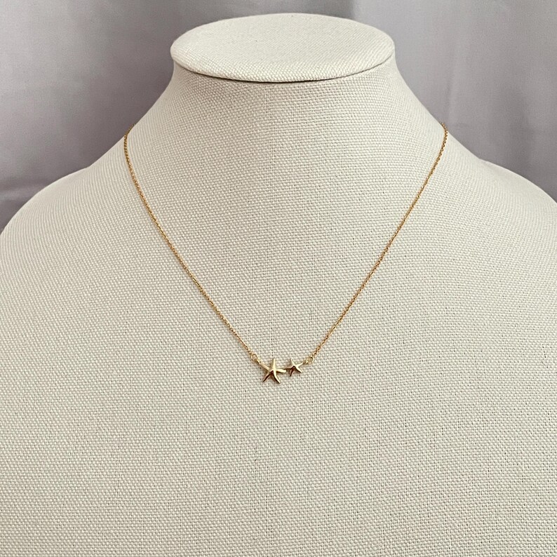 May include: A gold-toned necklace featuring a delicate chain and two small starfish-shaped pendants positioned together. The metal has a polished finish, and the design includes one larger starfish charm next to a slightly smaller starfish charm.