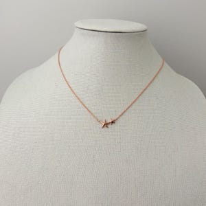 May include: A rose gold-toned necklace featuring a delicate chain and two small, five-pointed starfish charms positioned side-by-side at the center. The metallic finish has a warm, pinkish hue, and the chain is thin and simple in its design.