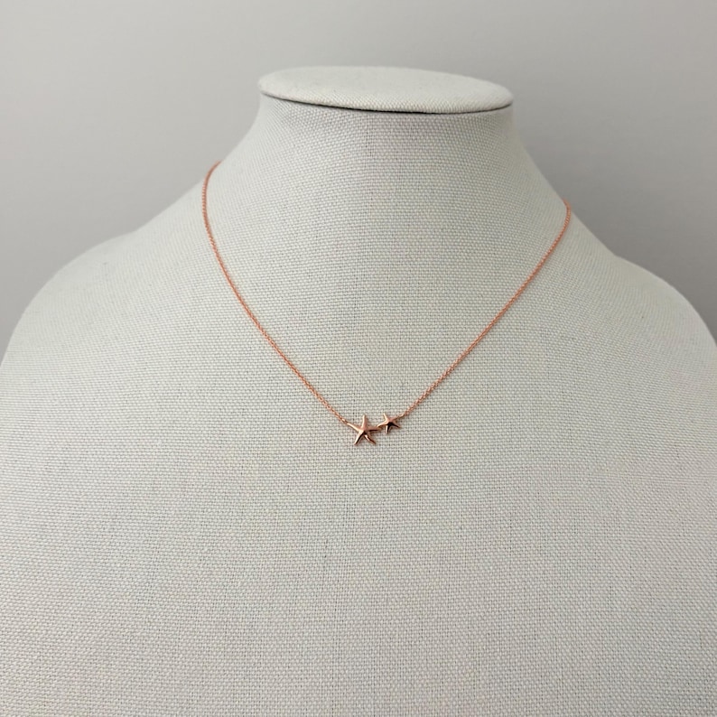 May include: A rose gold-toned necklace featuring a delicate chain and two small, five-pointed starfish charms positioned side-by-side at the center. The metallic finish has a warm, pinkish hue, and the chain is thin and simple in its design.