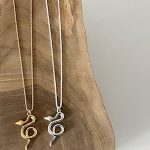Jorie Breonn Žaltys Snake Necklaces in gold and silver against a wood background.