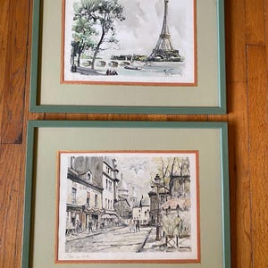 May include: Two framed watercolor prints of Paris scenes. The top print depicts the Eiffel Tower and the Seine River. The bottom print shows a bustling street scene with buildings and pedestrians. Both prints feature sage green frames with orange matting.
