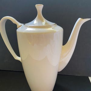 May include: A tall, off-white porcelain teapot with a smooth, iridescent finish. It features a slender, curved handle, a long, elegant spout, and a matching lid with a pointed finial. The design is clean and vertical, with gold-toned trim.