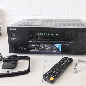 Może przedstawiać: Czarny amplituner AV Onkyo z cyfrowym wyświetlaczem pokazującym FM 89.1 MHz, z oznaczeniami Dolby Atmos i DTS:X. Urządzenie posiada duże pokrętło głośności, przycisk zasilania, gniazdo słuchawkowe, pilot zdalnego sterowania oraz ramową antenę.