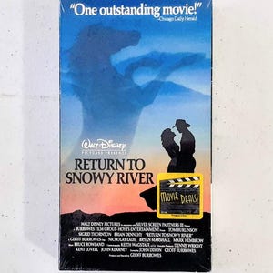 May include: A VHS tape case for the movie "Return to Snowy River" featuring a silhouette of a horse and a couple standing in front of a sunset. The text "One outstanding movie!" is printed on the top of the case. The text "WALT DISNEY PICTURES PRESENTS" is printed in a white box on the case. The text "RETURN TO SNOWY RIVER" is printed in a large font on the case. A yellow sticker with the text "MOVIE DEALS!" is on the bottom right corner of the case.
