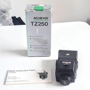 以下が含まれることがあります： 「Achiever」ブランド名とモデル番号「TZ250」が印刷された黒と銀のフラッシュユニット。フラッシュユニットには、箱と取扱説明書が付属しています。箱は緑色のアクセントがついた白で、ブランド名とモデル番号が記載されています。取扱説明書は「Multi-Dedicated Flash TZ250」というタイトルです。