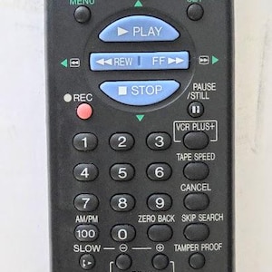 May include: A black Sharp remote control for a video cassette recorder with a red power button, blue playback control buttons, and a numeric keypad. The device features labels including VCR Plus+, Tape Speed, Skip Search, and model number RRMCG0120AJSA.
