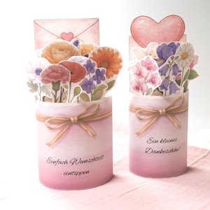May include: Two pink paper flower arrangements with a watercolor effect. One has an envelope topper, the other a heart. Each has a ribbon and text in German. The flowers are in shades of orange, red, purple, and white.