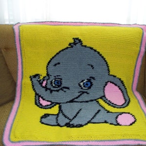Puede incluir: Una manta de beb&eacute; de crochet con fondo amarillo y borde rosa. La manta presenta un elefante gris con orejas rosas y ojos azules.
