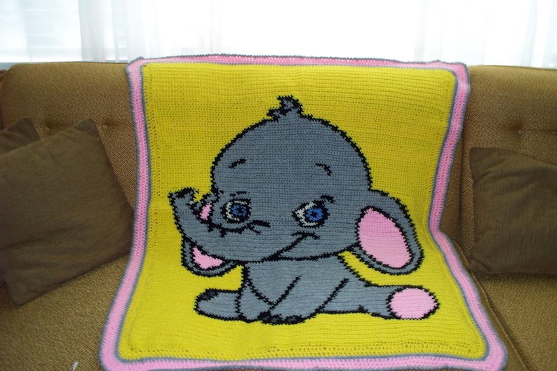 Puede incluir: Una manta de beb&eacute; de crochet con fondo amarillo y borde rosa. La manta presenta un elefante gris con orejas rosas y ojos azules.