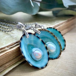 Könnte beinhalten: Drei türkisblaue Muschelketten mit silbernen Ketten und schillernden Perlen. Die Muscheln haben einen schwarzen Rand und sind übereinander gelegt.