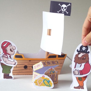 May include: A paper craft pirate ship with a white sail and a skull and crossbones flag. There are two paper pirate figures, one with a red bandana and one with a red shirt and a beard. A treasure chest with gold coins and jewels is in front of the ship.