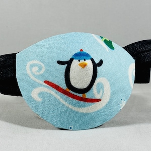May include: A blue eye patch with an elastic strap. The patch features a cartoon penguin wearing a blue hat and riding a surfboard on a wave.