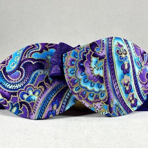 Pode incluir: Uma gravata borboleta de tecido com padr&atilde;o paisley roxo e azul com acabamento dourado. A gravata borboleta &eacute; feita de tecido e tem uma fita de cetim.