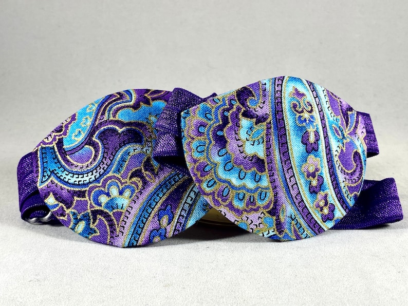 Pode incluir: Uma gravata borboleta de tecido com padr&atilde;o paisley roxo e azul com acabamento dourado. A gravata borboleta &eacute; feita de tecido e tem uma fita de cetim.