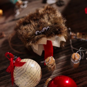 Puede incluir: Un adorno de monstruo peludo marr&oacute;n con ojos saltones y una lengua roja. El adorno est&aacute; rodeado de otras decoraciones navide&ntilde;as, incluyendo un adorno rojo y blanco, una pi&ntilde;a y una peque&ntilde;a botella de vidrio.