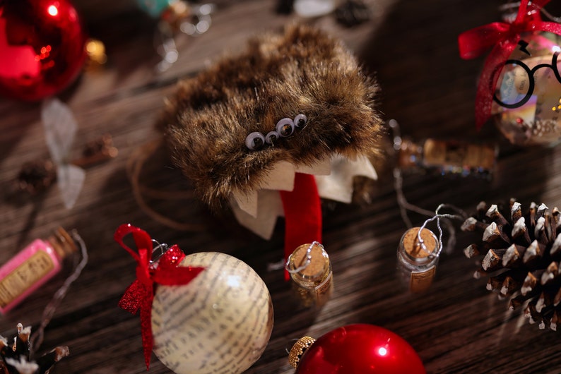 Puede incluir: Un adorno de monstruo peludo marr&oacute;n con ojos saltones y una lengua roja. El adorno est&aacute; rodeado de otras decoraciones navide&ntilde;as, incluyendo un adorno rojo y blanco, una pi&ntilde;a y una peque&ntilde;a botella de vidrio.