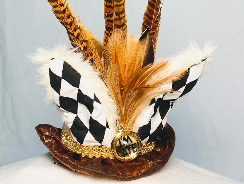 May include: A brown top hat with a black and white checkered band, gold trim, and a pocket watch with the words "I'm Late" on the face. The hat is decorated with brown and white feathers.