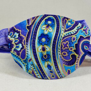 Pode incluir: Atadura para o olho roxa com um padr&atilde;o paisley em tons de azul, dourado e branco. A atadura tem uma faixa el&aacute;stica ajust&aacute;vel.
