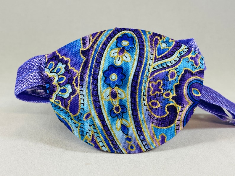 Pode incluir: Atadura para o olho roxa com um padr&atilde;o paisley em tons de azul, dourado e branco. A atadura tem uma faixa el&aacute;stica ajust&aacute;vel.