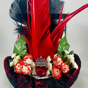 May include: A black and red top hat with a red heart, a silver crown, and red and white roses. The hat is decorated with black and red feathers.
