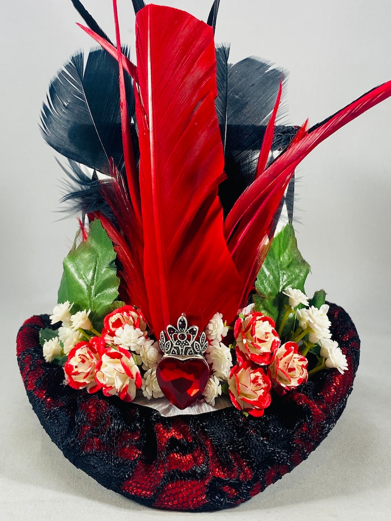 May include: A black and red top hat with a red heart, a silver crown, and red and white roses. The hat is decorated with black and red feathers.