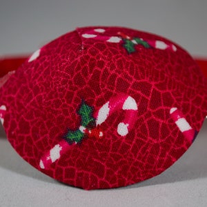 May include: A red eye patch with a white and green candy cane and holly pattern. The eye patch is made of fabric and has a red elastic band.