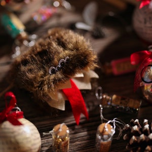 Puede incluir: Un adorno de piel marr&oacute;n con ojos saltones y una cinta roja. El adorno cuelga de una cadena plateada. Hay otros adornos navide&ntilde;os en el fondo, incluyendo un adorno de vidrio transparente con un par de gafas dentro.