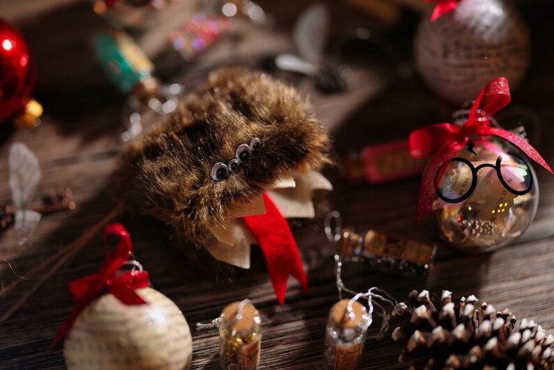 Puede incluir: Un adorno de piel marr&oacute;n con ojos saltones y una cinta roja. El adorno cuelga de una cadena plateada. Hay otros adornos navide&ntilde;os en el fondo, incluyendo un adorno de vidrio transparente con un par de gafas dentro.