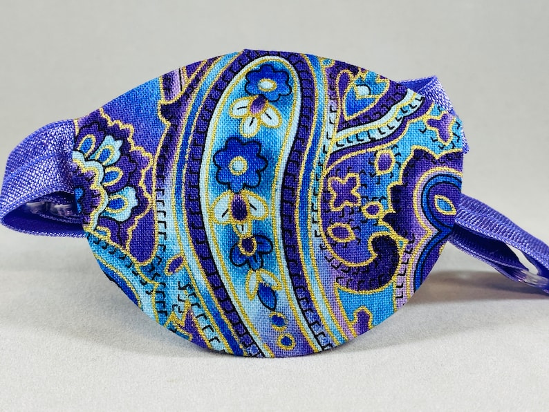 Pode incluir: Atadura para o olho roxa com um padr&atilde;o paisley em tons de azul, dourado e roxo. A atadura tem uma al&ccedil;a ajust&aacute;vel.
