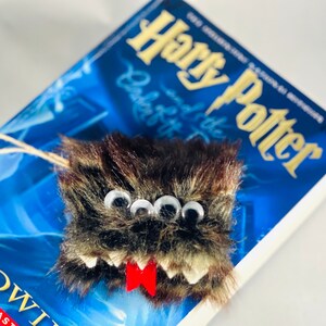 Puede incluir: Un marcap&aacute;ginas de monstruo peludo marr&oacute;n y negro con tres ojos saltones y una lengua roja. El marcap&aacute;ginas est&aacute; encima de un libro de Harry Potter.
