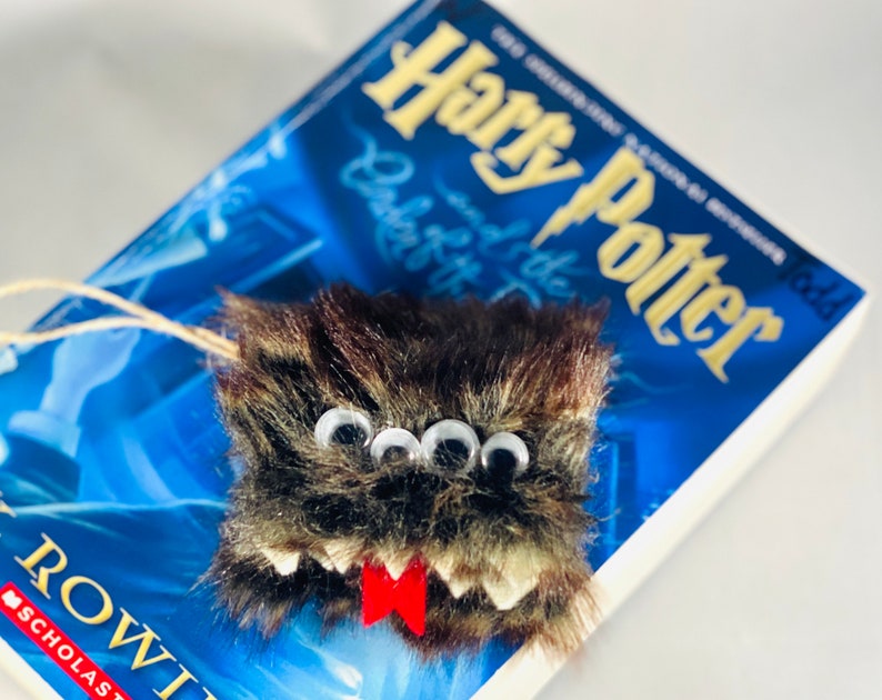 Puede incluir: Un marcap&aacute;ginas de monstruo peludo marr&oacute;n y negro con tres ojos saltones y una lengua roja. El marcap&aacute;ginas est&aacute; encima de un libro de Harry Potter.