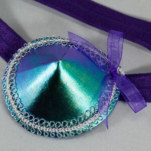 Peut inclure: Une paire de pasties bleu et vert irisés avec des bordures argentées et un ruban violet. Les pasties sont attachés à une sangle élastique violette.