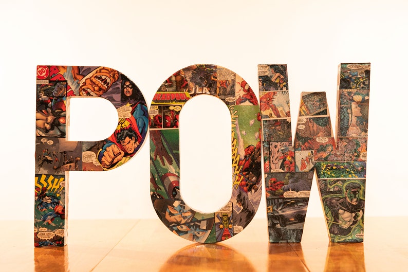 Op de afbeelding: Het woord "POW" is gespeld in grote letters gemaakt van karton bedekt met comicboekpagina's. De letters zijn kleurrijk en bevatten afbeeldingen van superhelden en schurken.