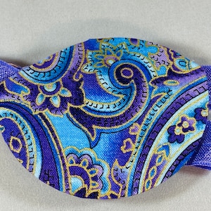 Pode incluir: Atadura para o olho roxa e azul com um padr&atilde;o paisley e detalhes dourados. A atadura tem uma faixa el&aacute;stica.
