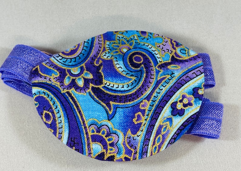 Pode incluir: Atadura para o olho roxa e azul com um padr&atilde;o paisley e detalhes dourados. A atadura tem uma faixa el&aacute;stica.