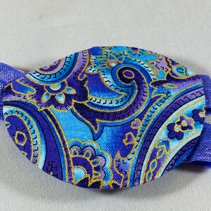 Pode incluir: Um tapa-olho azul e roxo com um padr&atilde;o paisley e detalhes dourados. O tapa-olho est&aacute; preso a uma faixa el&aacute;stica azul.